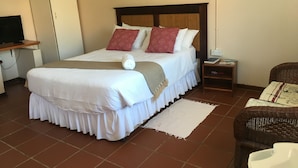 Una habitación de hotel ordenada con una cama grande, una mesita de noche con lámpara, un televisor sobre un soporte y una silla de mimbre.