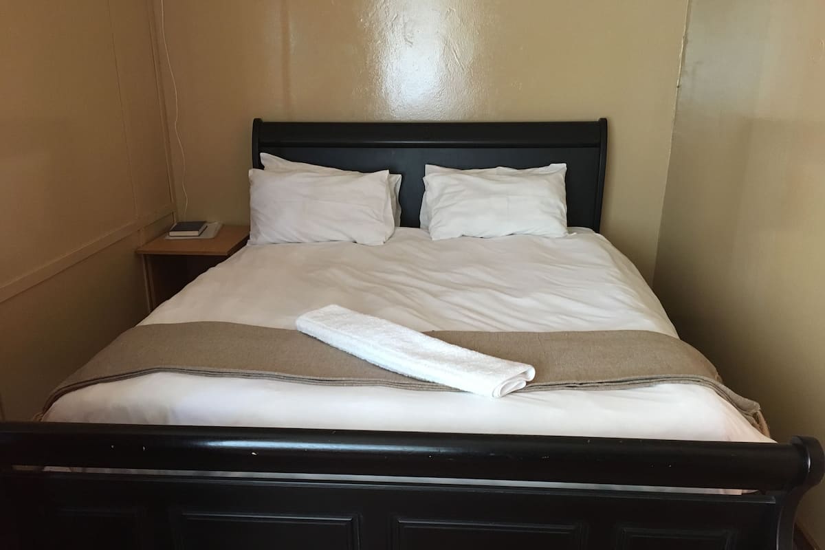 A neatly made bed with white linens and a folded towel, a bedside table with a book and a phone, and a dark wooden headboard.