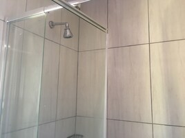 Una ducha con mampara de vidrio, cabezal de ducha de mano, paredes de azulejos y una esquina visible.