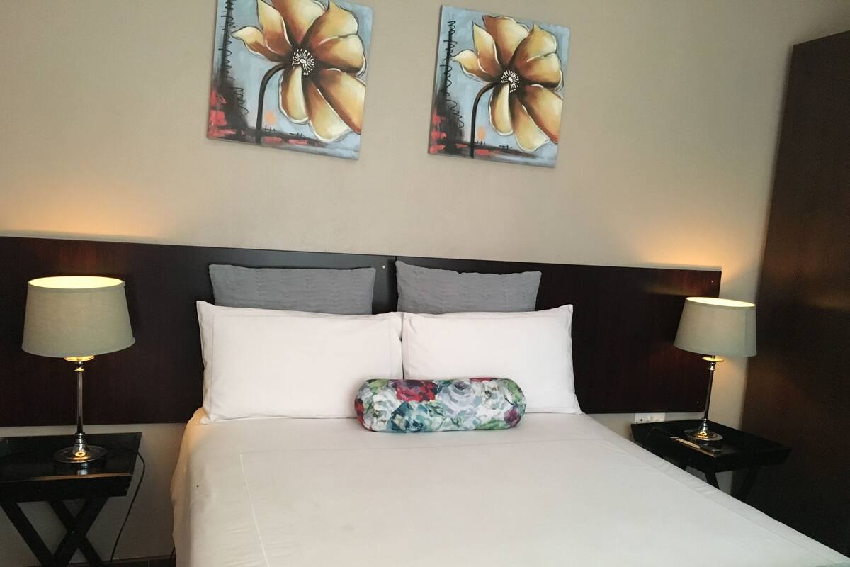 A hotel room with a large bed, two bedside lamps, a floral pillow, and two wall-mounted flower paintings.
