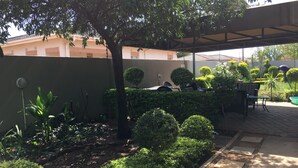 A garden area with a covered seating area, neatly trimmed bushes, and a tree providing shade. - Yana Bed & Breakfast (Gaborone)