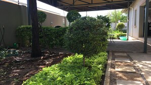A garden area with a paved walkway, a covered seating area, and a variety of plants and trees. - Yana Bed & Breakfast (Gaborone)