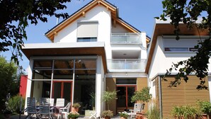 Une maison moderne à deux étages, dotée d’un balcon, d’un espace extérieur aménagé pour se détendre et de plantes en pot.