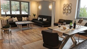 Un salon moderne avec un canapé, une table basse et un coin repas. On peut voir deux verres à vin et des bouteilles sur la table du coin repas.