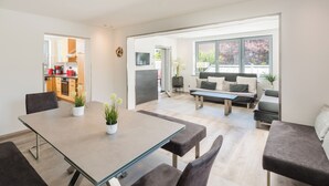 Un salon moderne avec une table à manger au plateau en verre, des canapés sombres et une grande fenêtre donnant sur l’extérieur.
