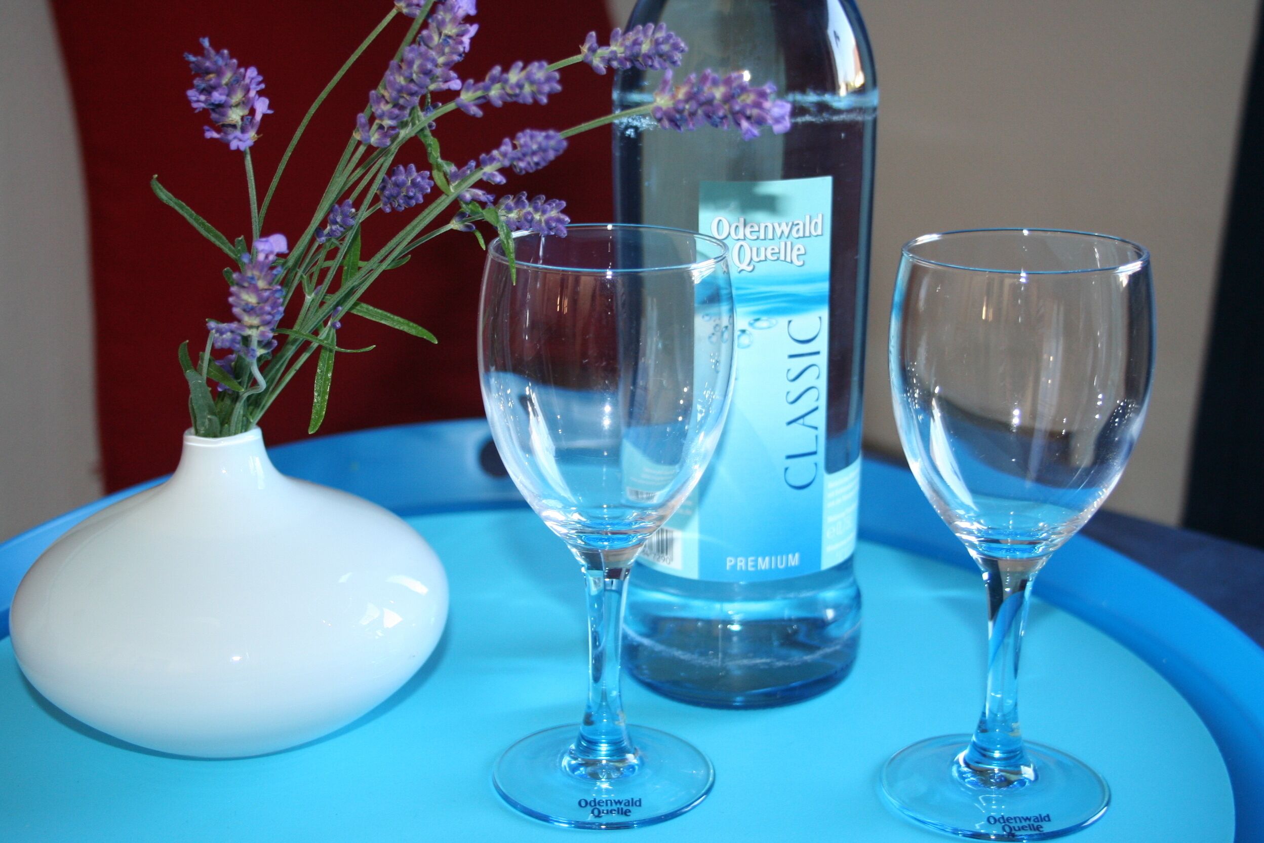 A bottle of Odenwald Quelle Classic water, two empty wine glasses, and a white vase with lavender flowers on a blue table.