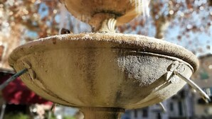 A close-up of a stone fountain with a metal spout, set against a blurred background of trees and buildings. - - Large cozy studio ---- (Puget-Théniers)