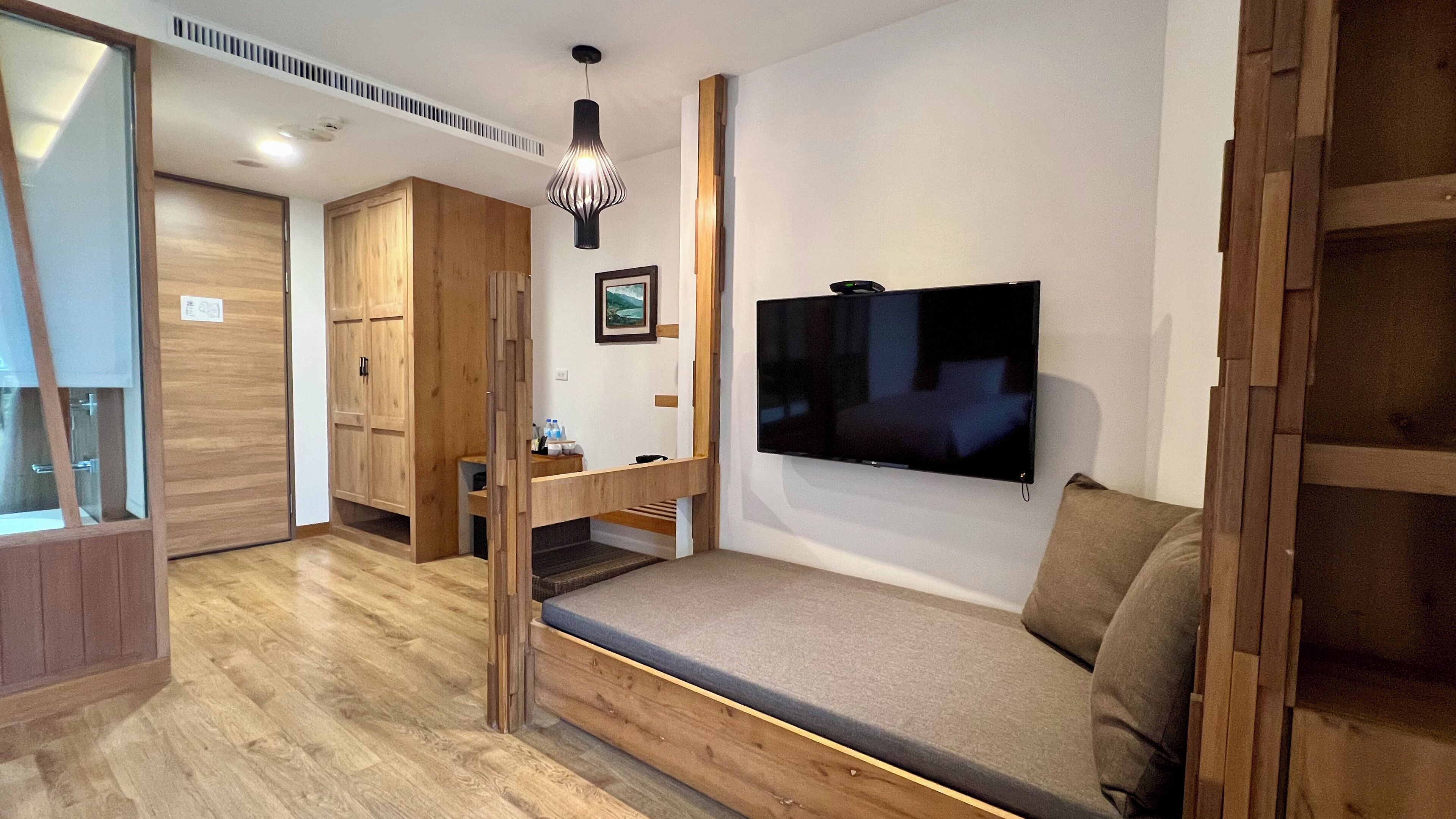 A modern room with a wooden floor, a built-in bench with cushions, a flat-screen TV mounted on the wall, and a small table with a picture frame and a few items on it.