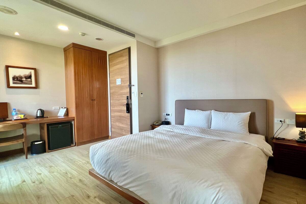 Una habitación de hotel con una cama grande, un escritorio con una silla, un armario y una mesita de noche con una lámpara.