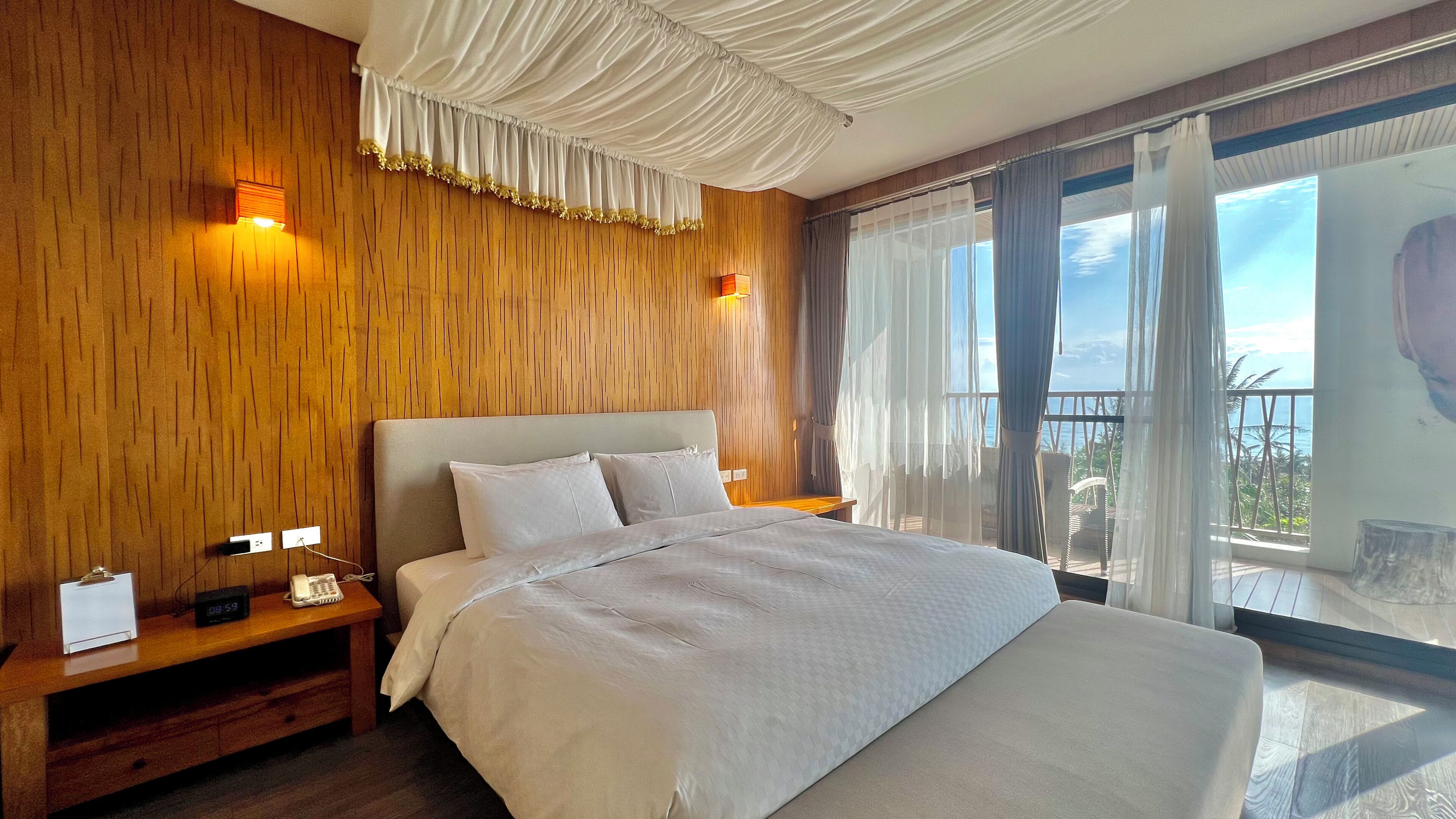 A hotel room with a large bed, wooden panel walls, a balcony with a view, and a clock on the bedside table.