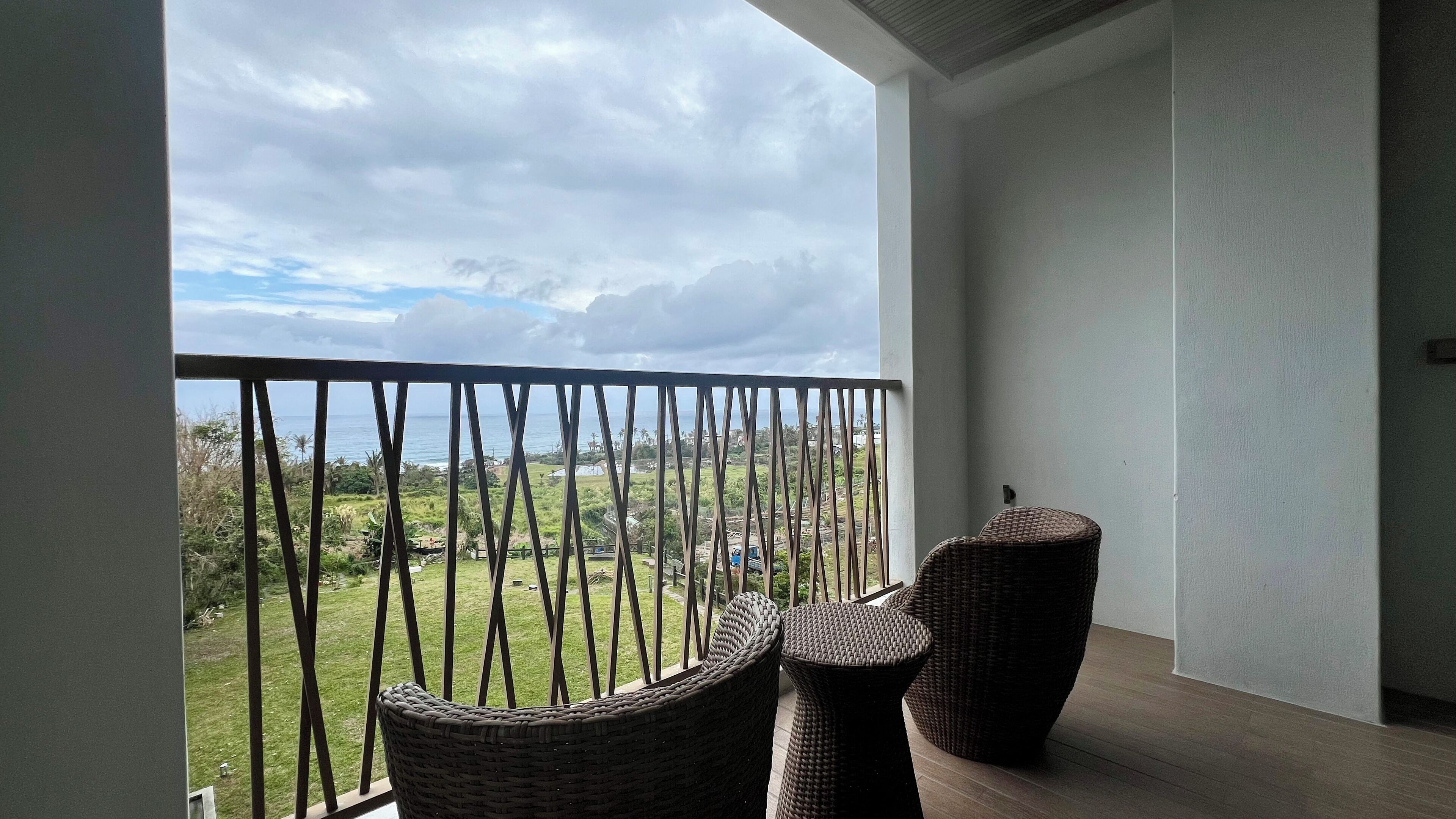 A balcony with a view of a green landscape and the sea. There are two wicker chairs and a small round table.