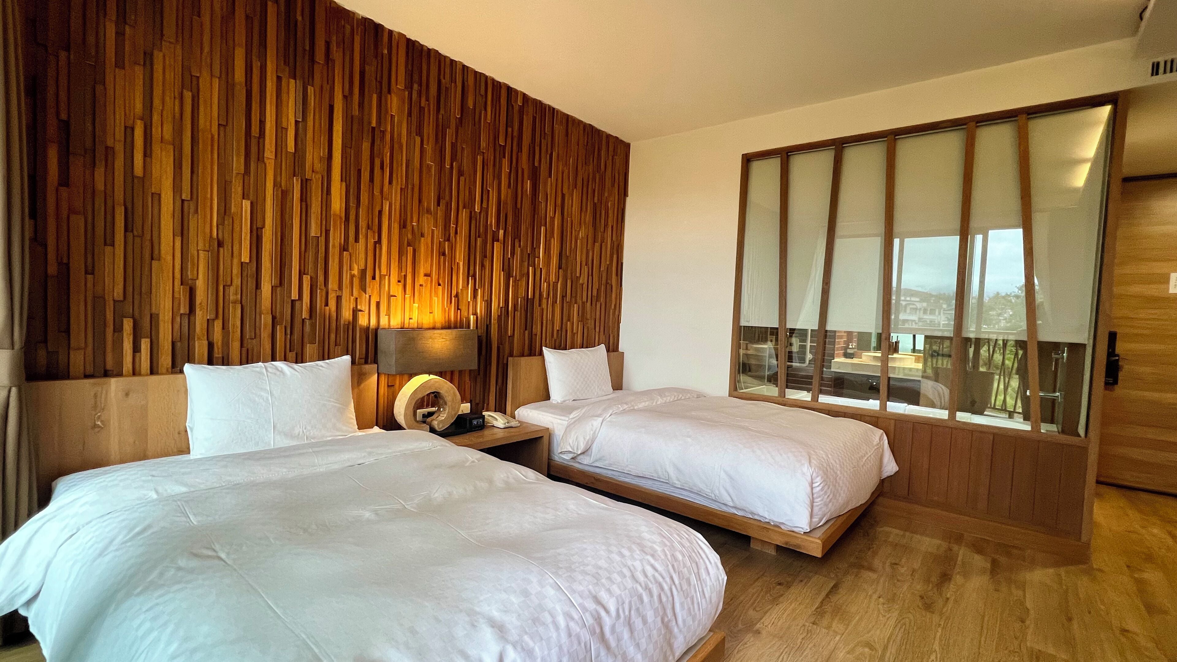 A hotel room with two beds, a wooden wall panel, a large window, and a wooden floor.