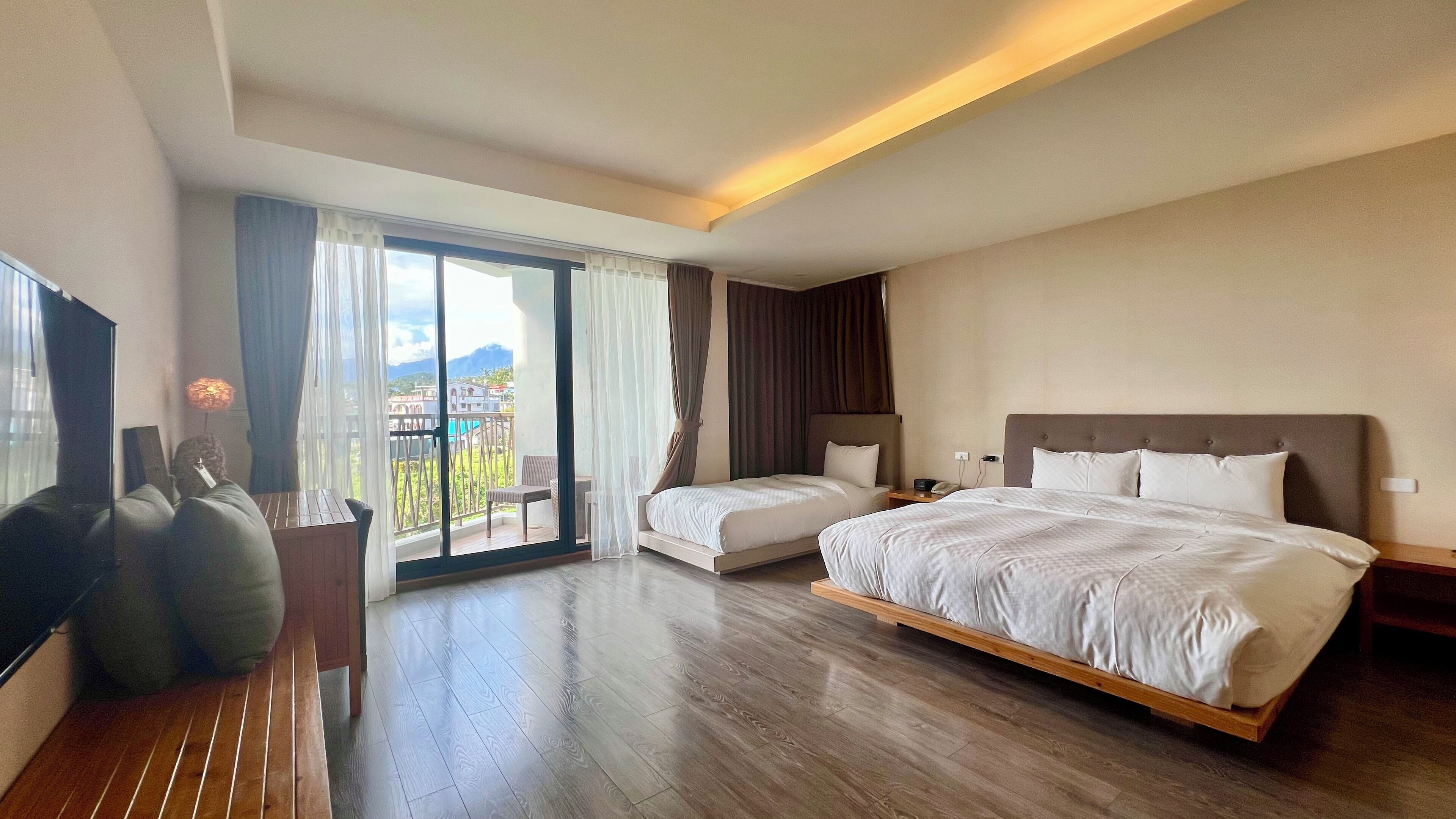 A hotel room with a large bed, a smaller bed, a sofa, a TV, and a balcony with curtains.