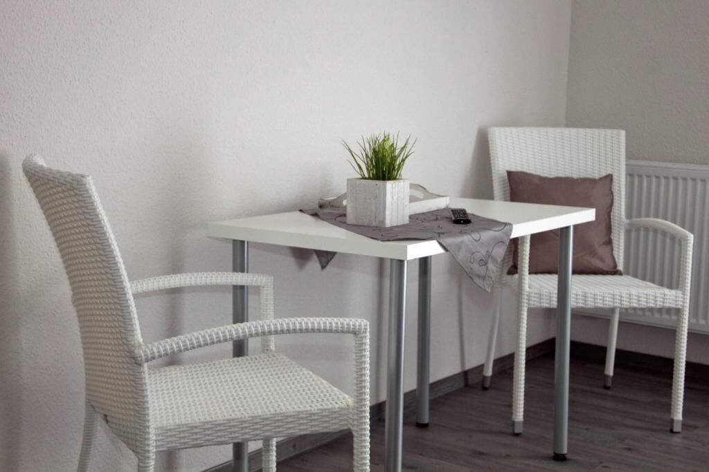 Ein kleines, weißes Rattangebäude mit Tisch und Stuhl vor einer schlichten Wand mit Heizkörper.