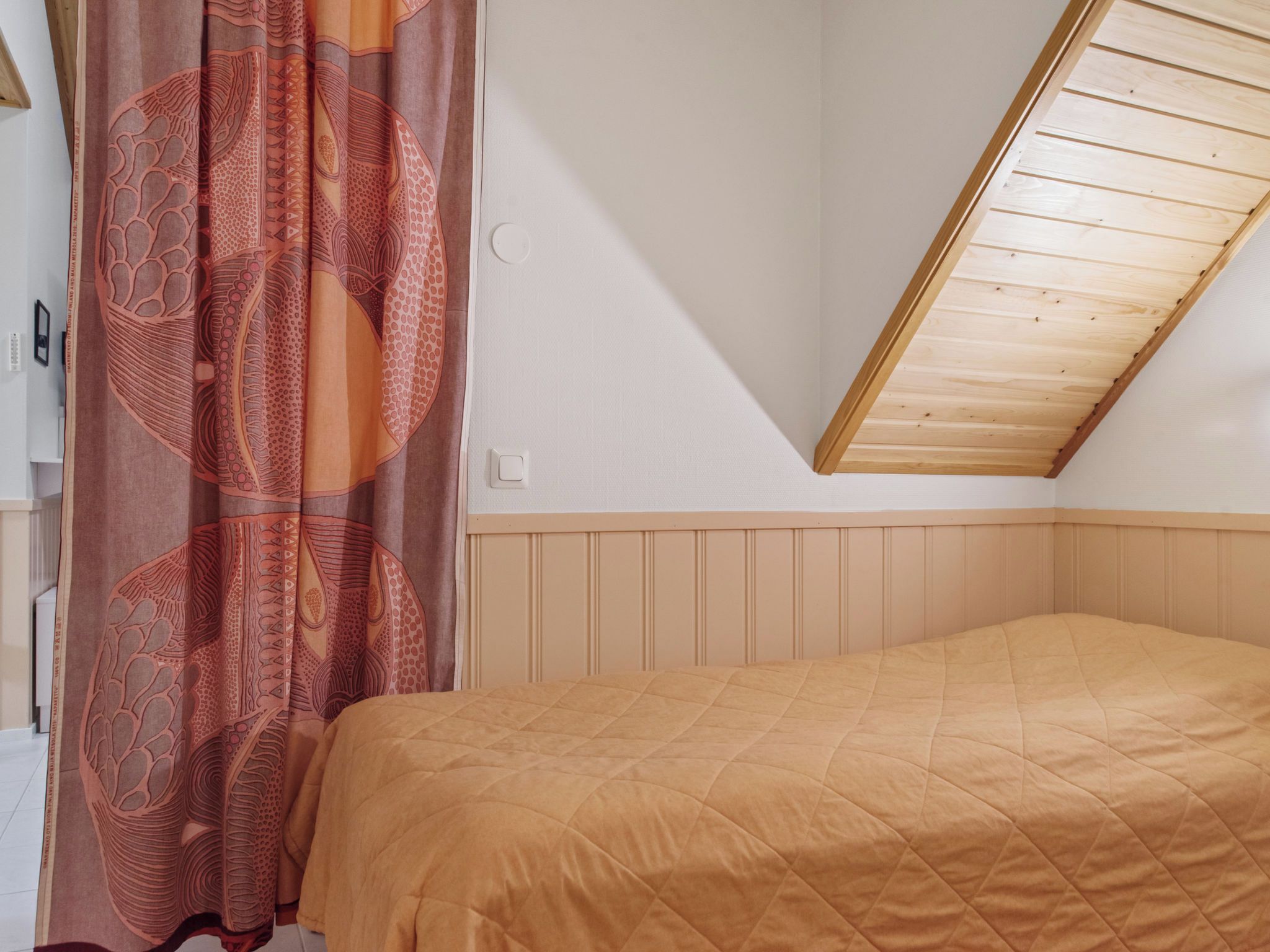 Une chambre avec un plafond en pente, un lit recouvert d’une courtepointe matelassée et un rideau à motifs.