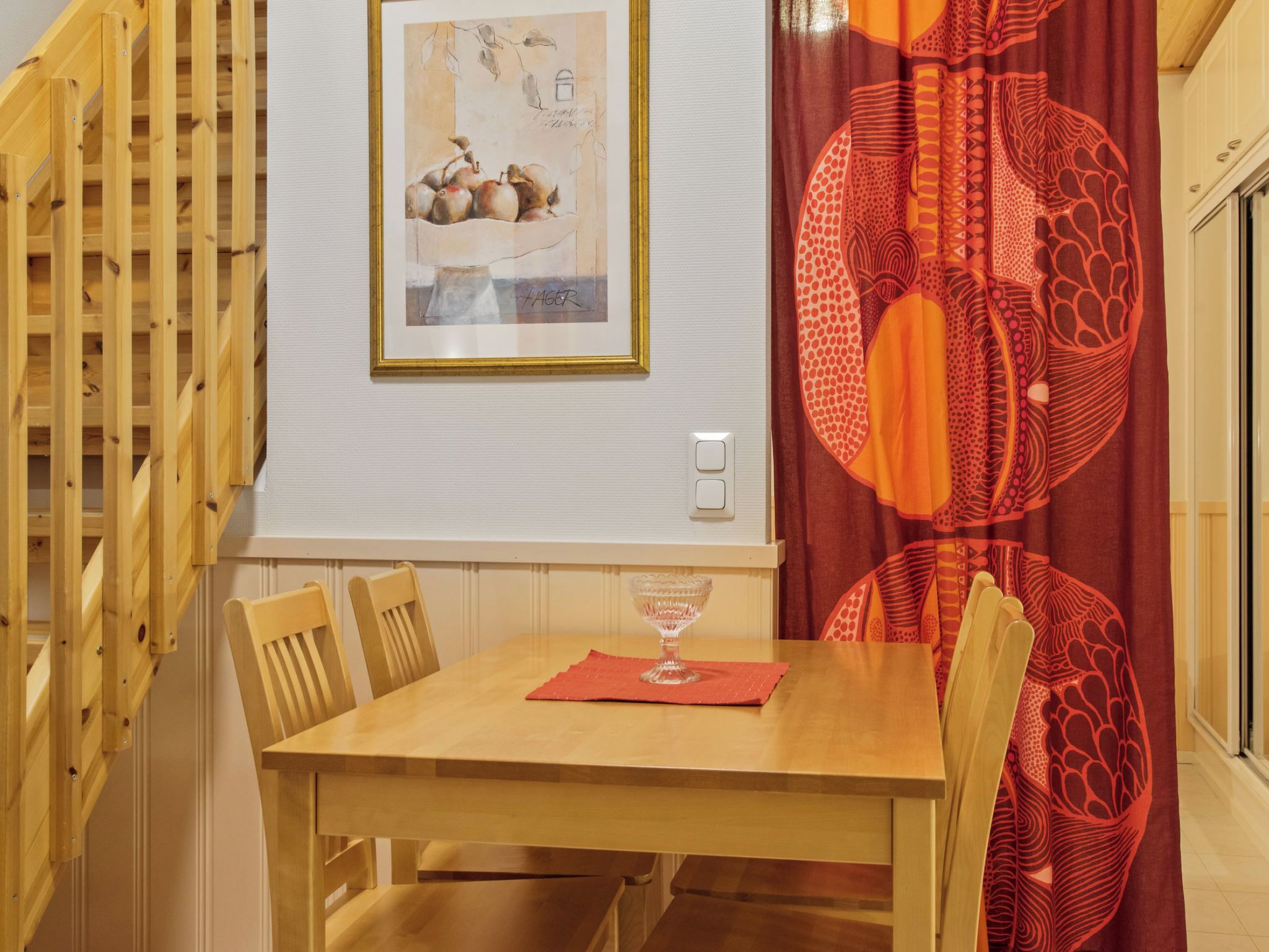Un coin repas avec une table et des chaises en bois, une œuvre d’art encadrée au-dessus de la table et un rideau rouge à motifs.