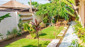Um jardim com um caminho pavimentado, diversas plantas e uma estrutura com telhado de palha.