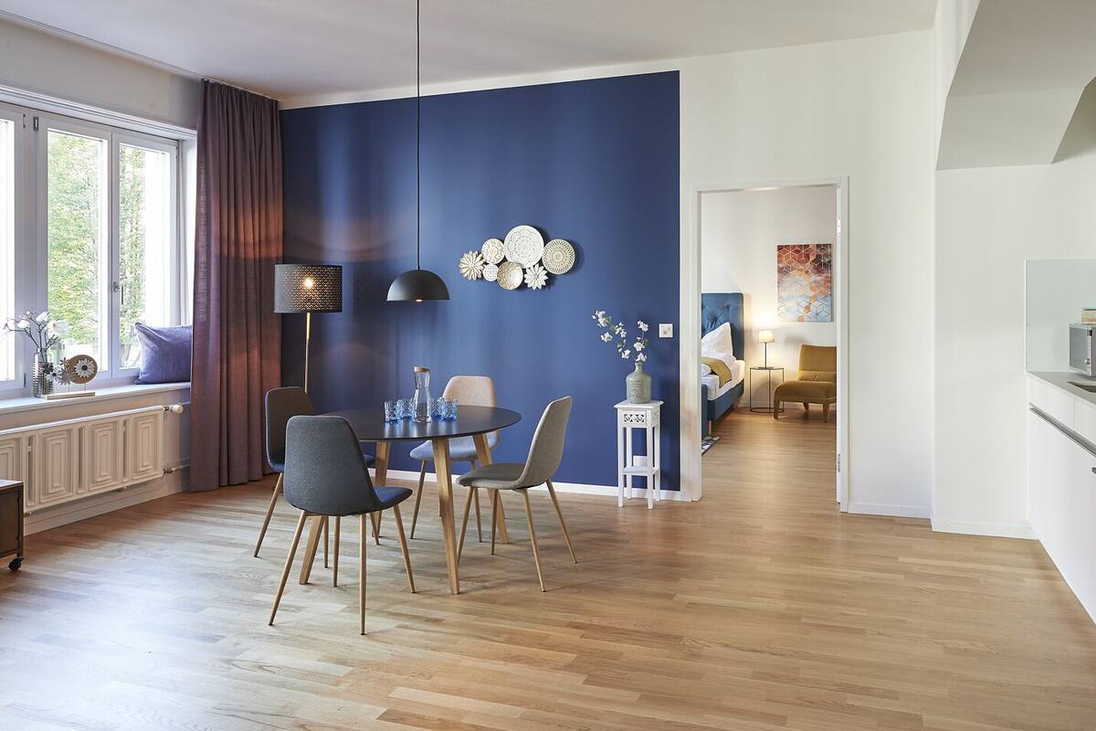 A modern living room with a dining area, featuring a blue accent wall, wooden flooring, and a view into the adjacent bedroom.