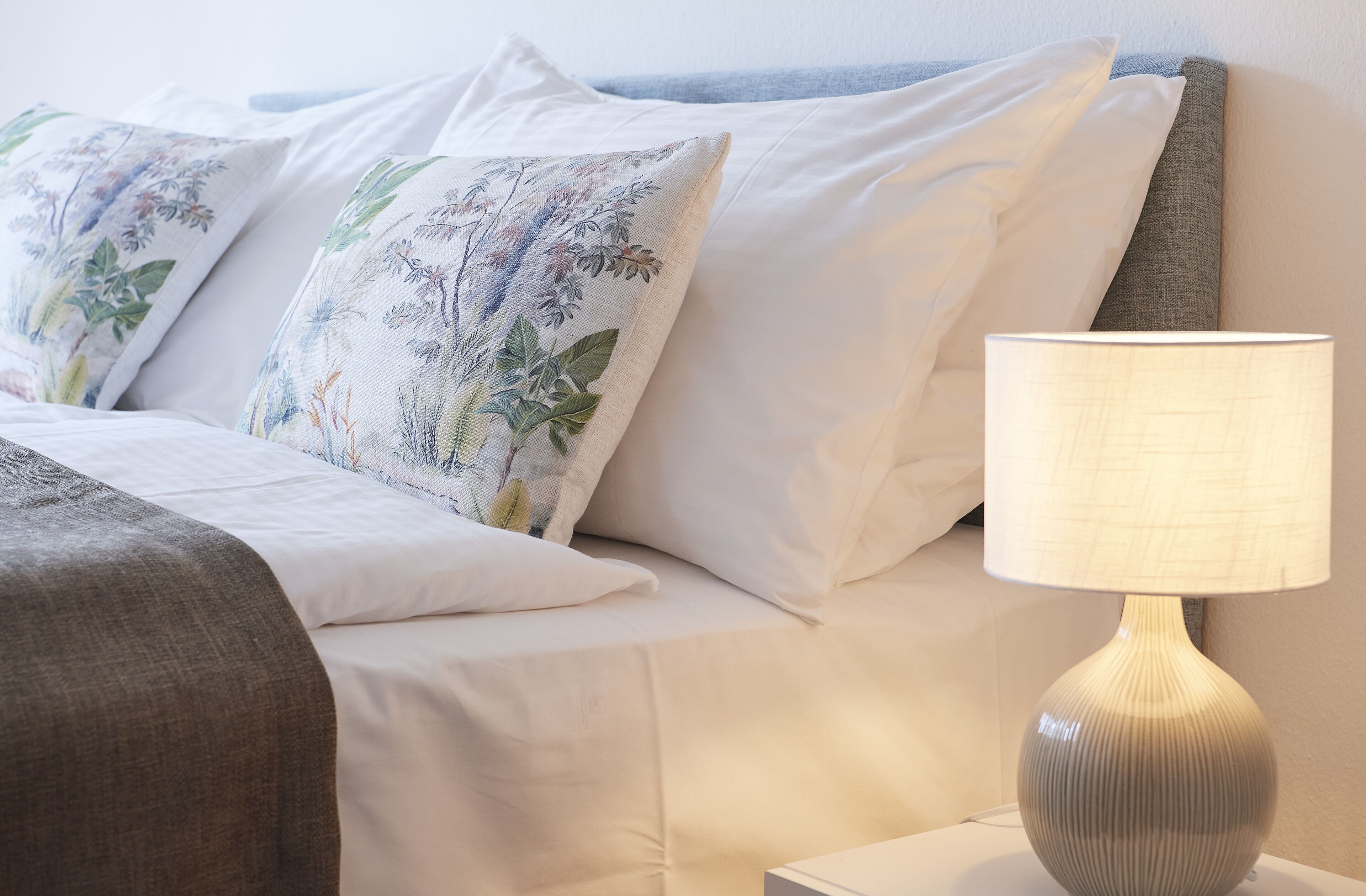 A neatly made bed with floral pillows, a grey throw blanket, and a table lamp.