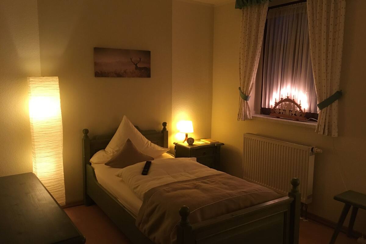 A bedroom with a bed, bedside lamps, a window with curtains, a radiator, and a wall-mounted picture.