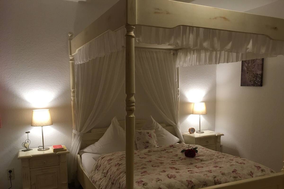 A four-poster bed with a floral bedspread and a red rose. There are two bedside tables with lamps and a clock.