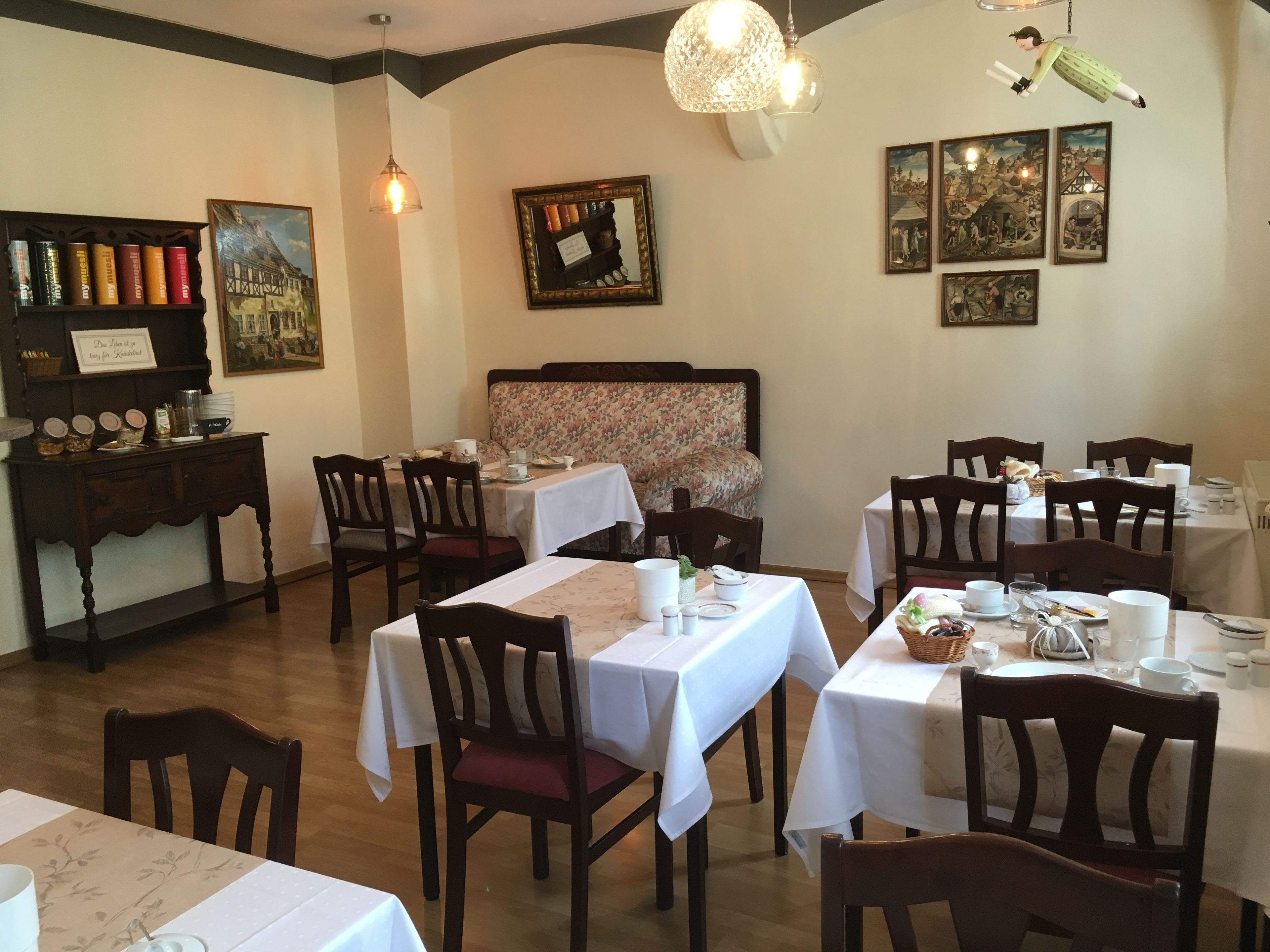 A dining area with tables set for service, wooden chairs, a floral sofa, and a wall with framed pictures.