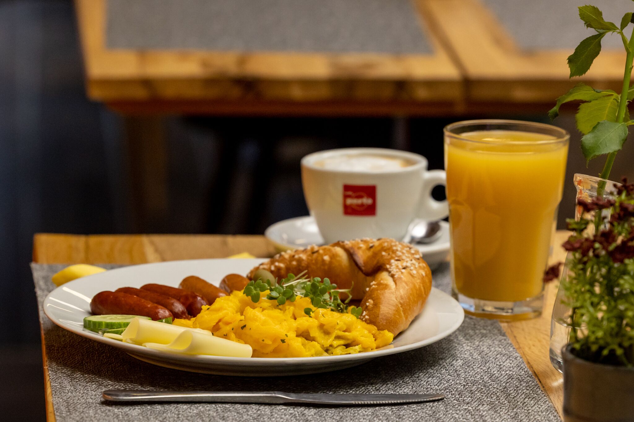 Un plato de desayuno con salchichas, huevos revueltos y un croissant, acompañado de una taza de café y un vaso de jugo de naranja.