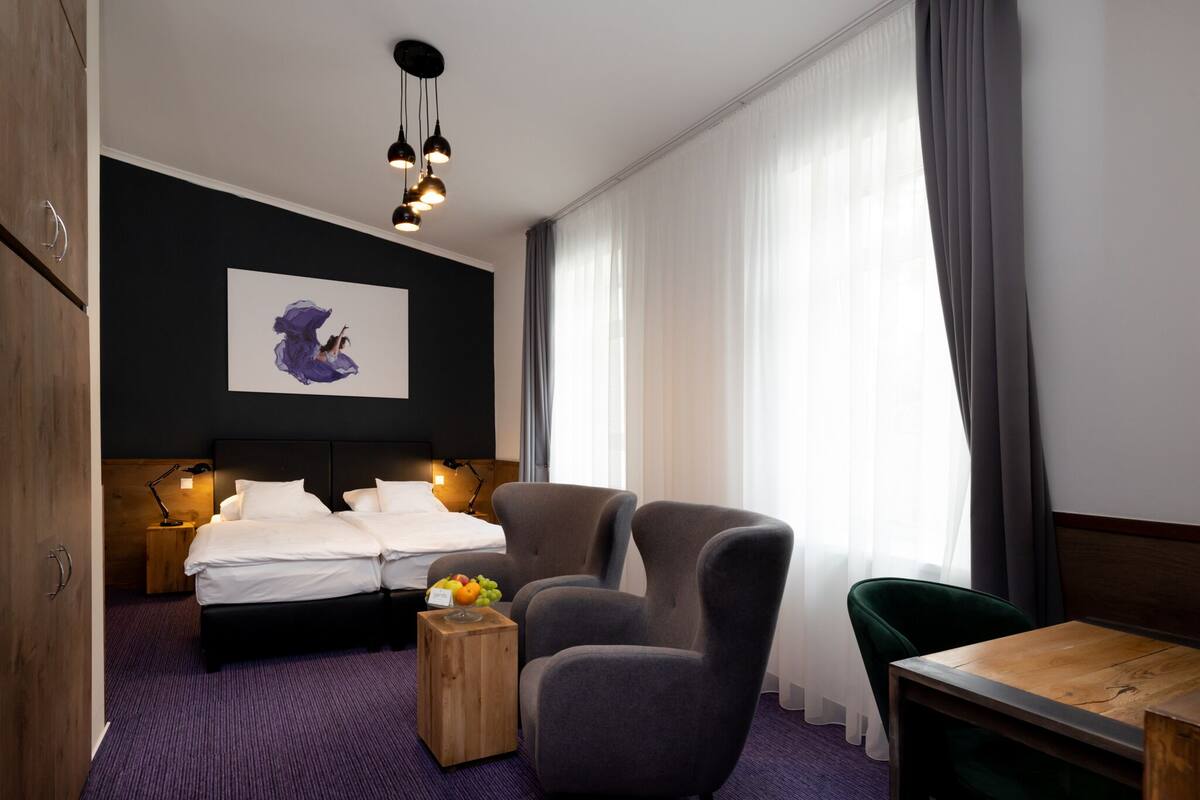 A hotel room with a bed, two armchairs, a wooden side table with fruit, and a painting on the wall.