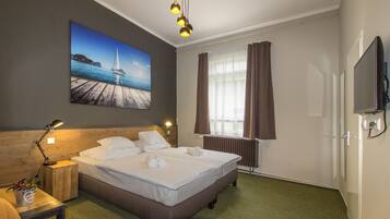 Una habitación de hotel con cama, mesita de noche, lámpara, un cuadro de un muelle, ventana con cortinas y televisor.
