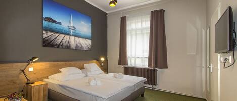 Una habitación de hotel con cama, mesita de noche, lámpara, un cuadro de un muelle, ventana con cortinas y televisor.
