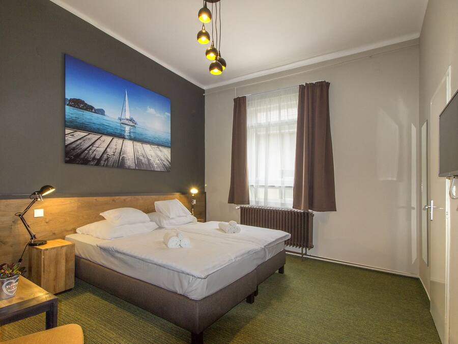Una habitación de hotel con cama, mesita de noche, lámpara, un cuadro de un muelle, ventana con cortinas y televisor.