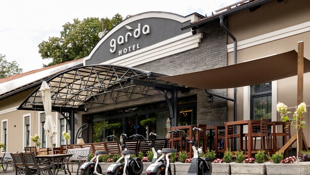 A hotel entrance with a covered patio area, outdoor seating, and electric scooters parked in front. - Garda Hotel (Szombathely)