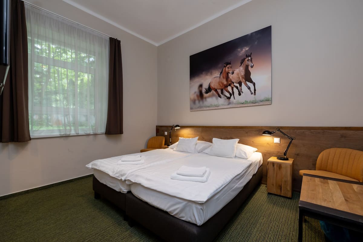 A hotel room with a large bed, two bedside tables, a chair, a painting of horses, and a window with curtains.
