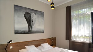 A hotel room with a large bed, two bedside tables, a headboard, a window with curtains, and a wall-mounted elephant painting. - Garda Hotel (Szombathely)