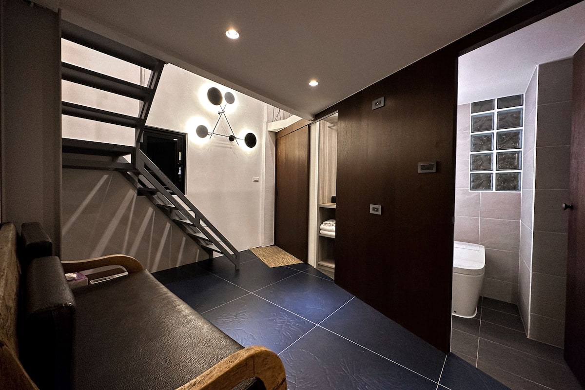 A modern bathroom with a shower, toilet, and a sink. There is a wooden bench and a staircase leading to another room.