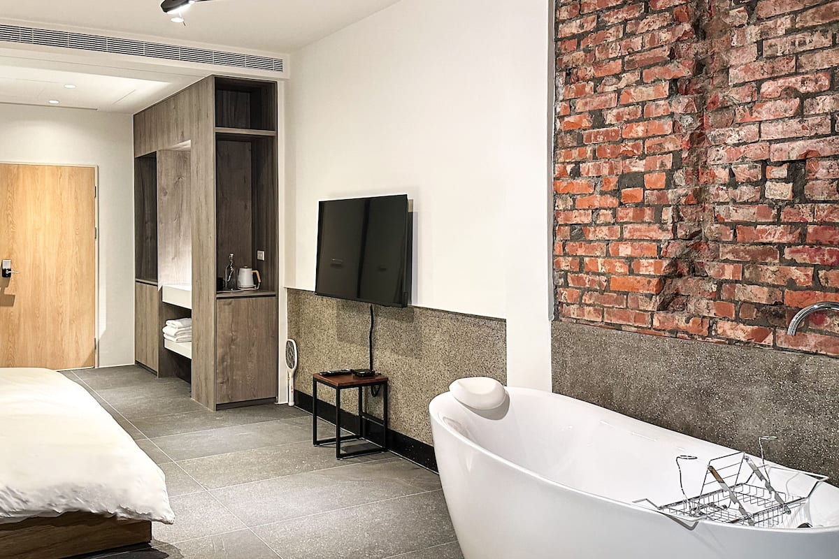 A modern bathroom with a freestanding bathtub, a wooden wardrobe, a flat-screen TV mounted on the wall, and a brick accent wall.