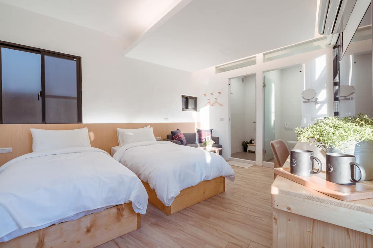 A modern hotel room with two beds, a wooden headboard, a small seating area with a chair and a table, and a potted plant.