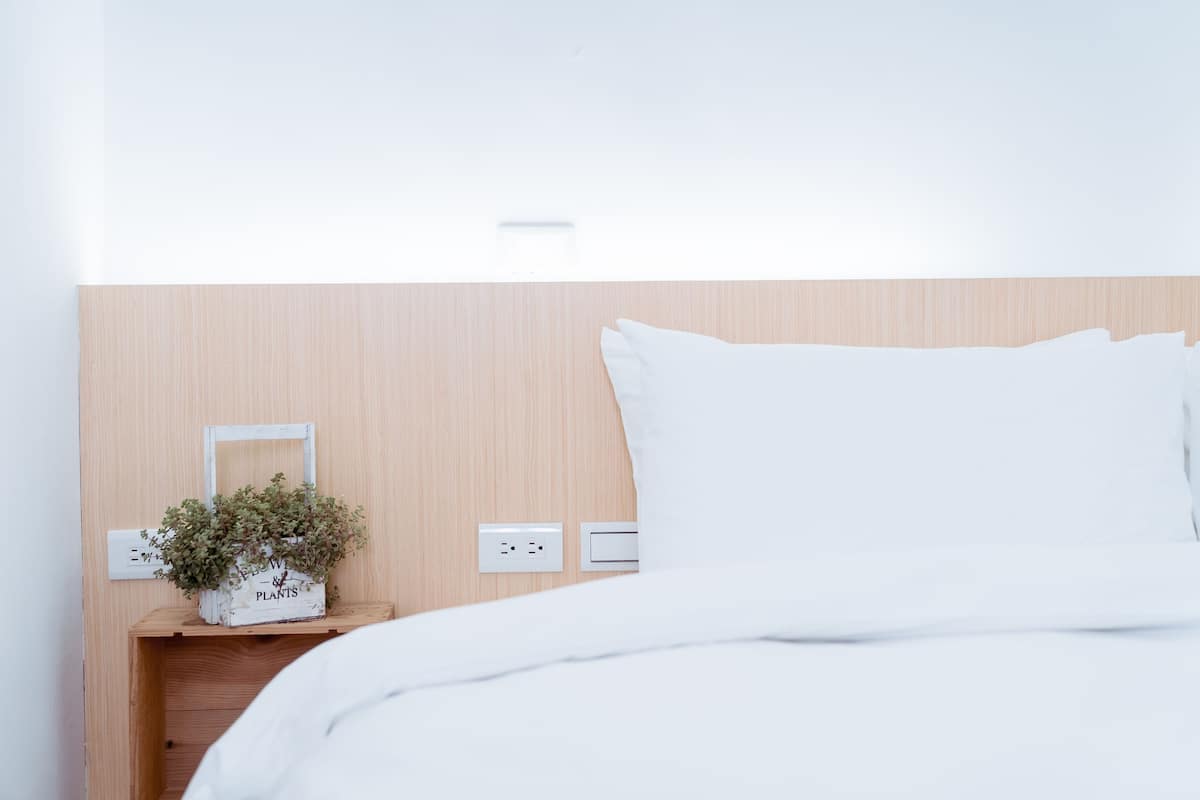 A neatly made bed with white bedding, a wooden headboard, a small wooden nightstand with a plant and a framed picture, and a wall socket.