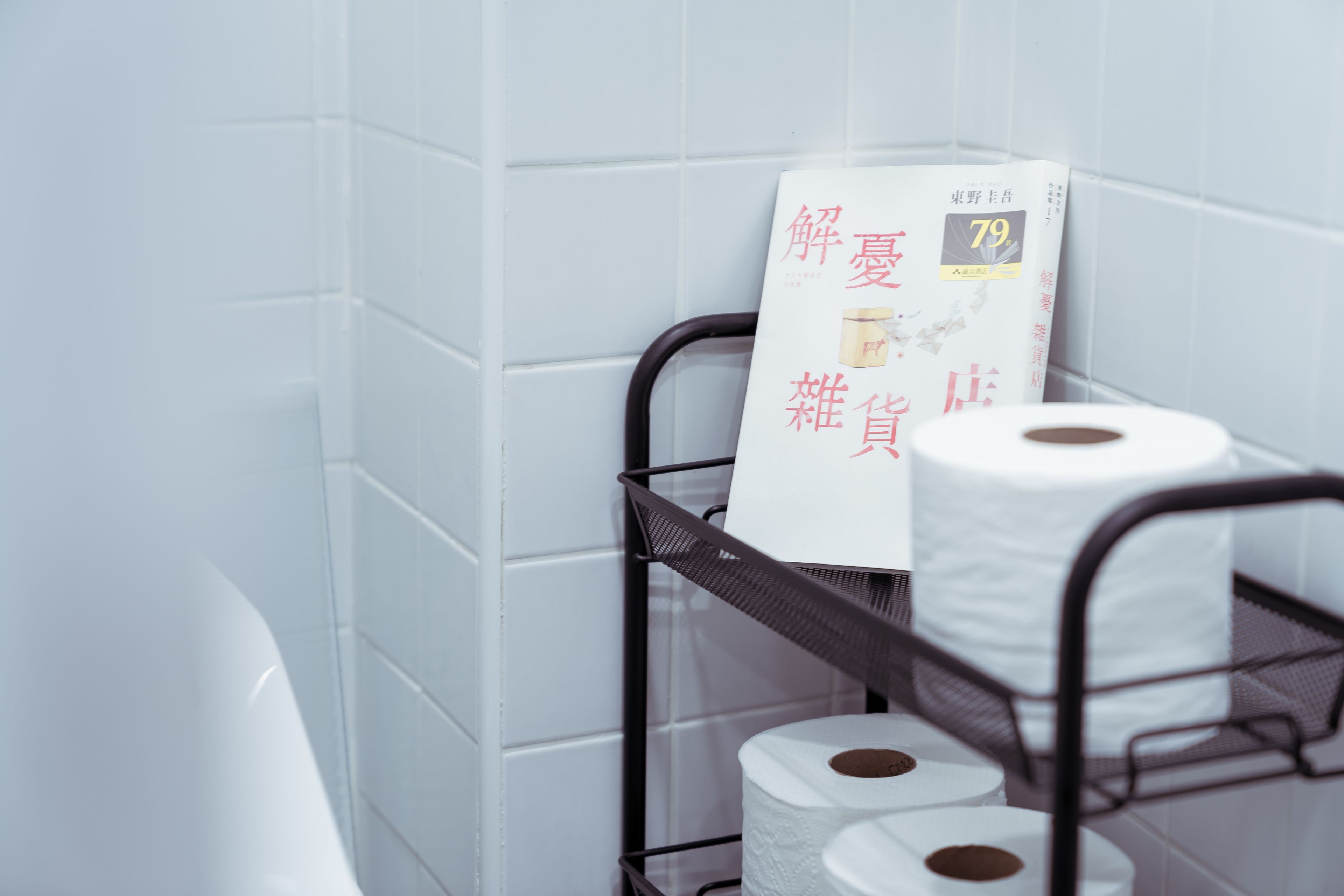 A bathroom with white tiles, a metal rack holding toilet paper and a magazine.