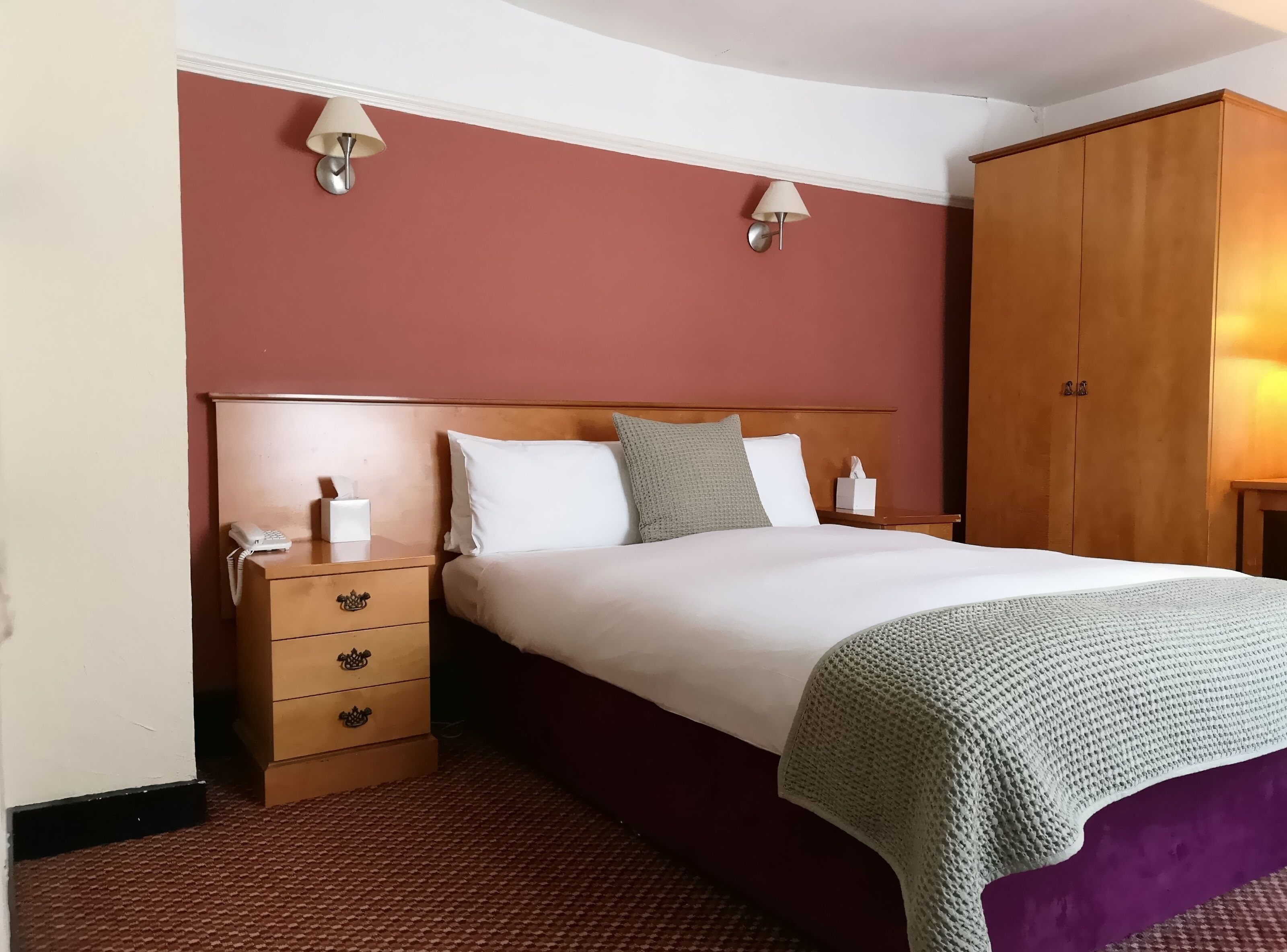 A hotel room with a wooden headboard, a bed with white bedding, two bedside lamps, a wooden wardrobe, and a telephone on the nightstand.