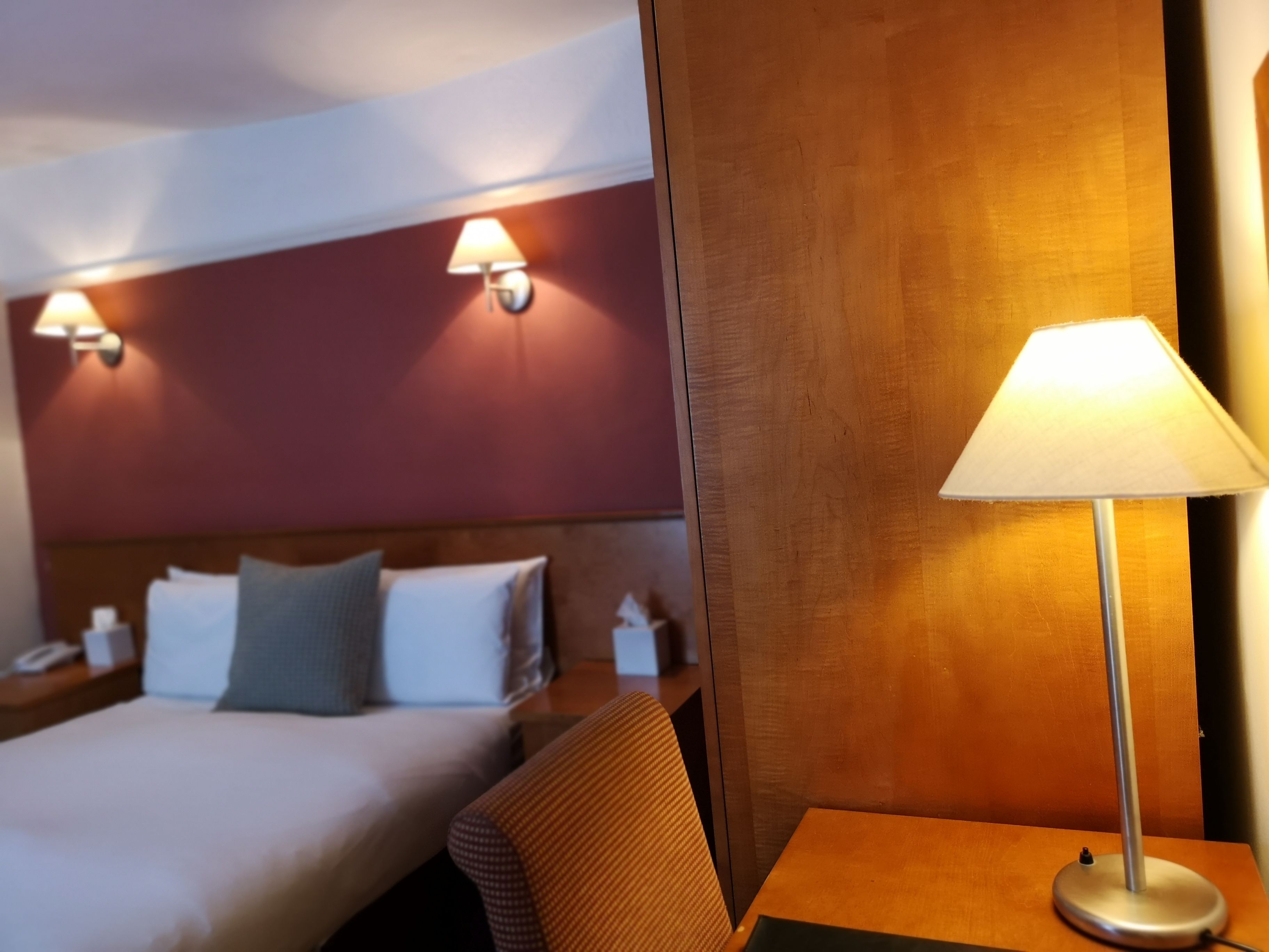A hotel room with a bed, a nightstand, a lamp, and a wall-mounted light fixture.