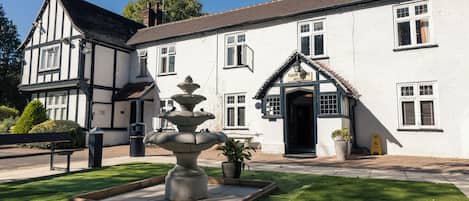 A Tudor Lodge Hotel with a stone fountain, a well-maintained lawn, and a traditional building facade.