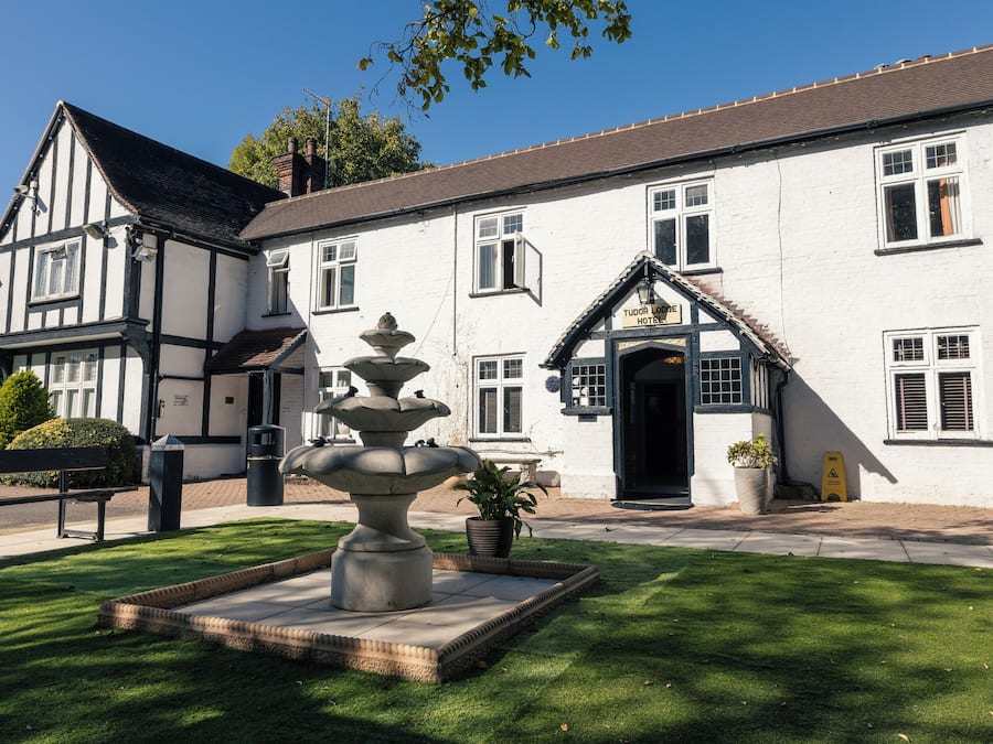 Hotel Tudor Lodge com uma fonte de pedra, um jardim bem cuidado e fachada de edifício tradicional.