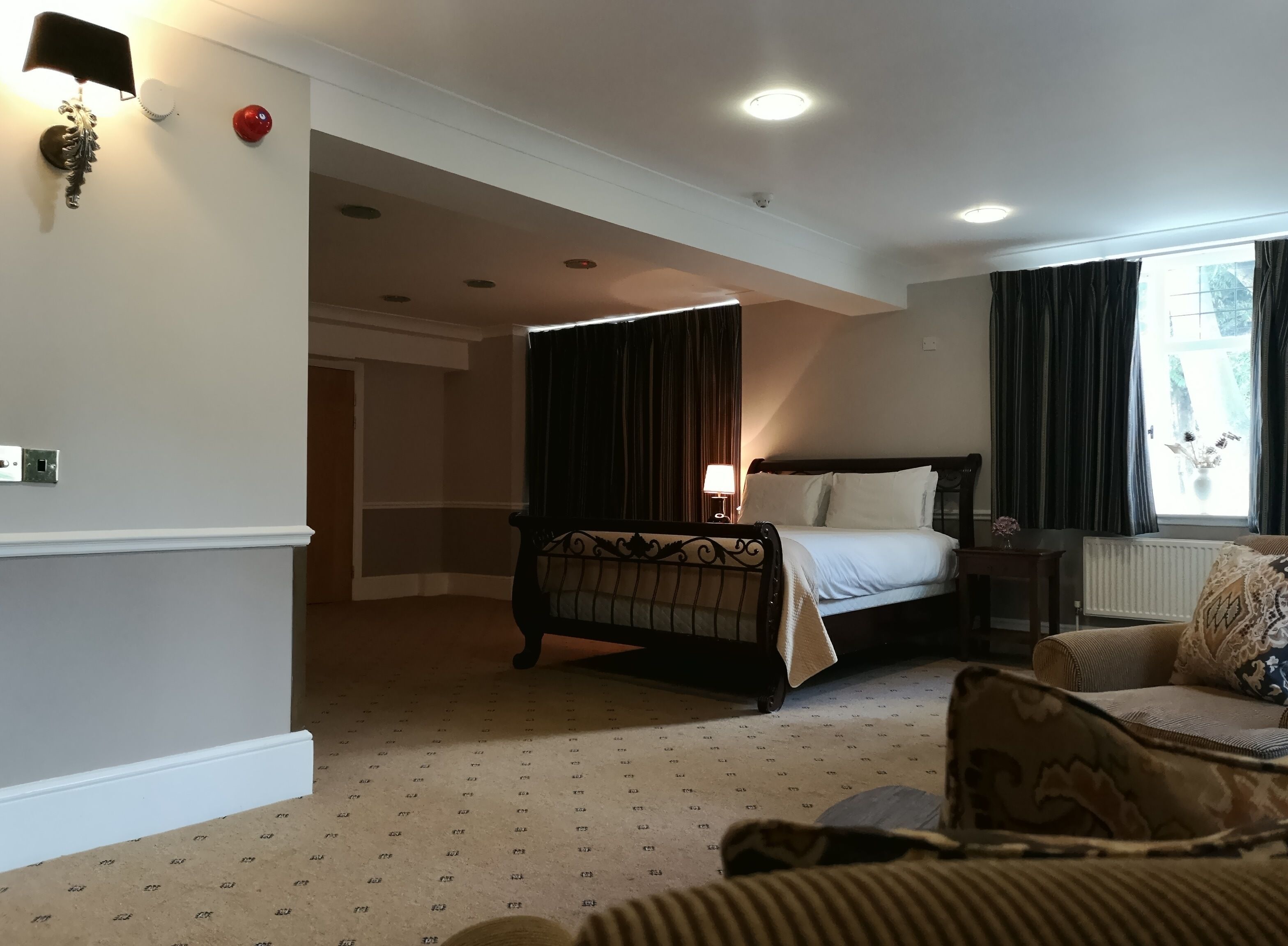 A hotel room with a bed, a chair, a lamp, a window with curtains, and a wall-mounted light fixture.