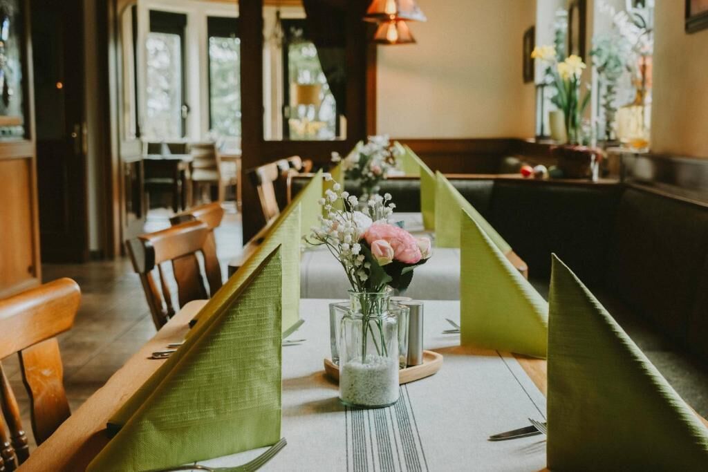 Ein Restaurantambiente mit einem für zwei Personen gedecktem Tisch, mit grünen Tischläufern, einer Blumenvase und einem Kerzenständer.