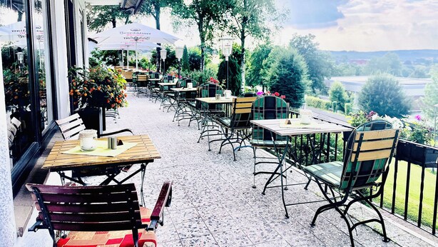 An outdoor cafe with tables and chairs, a view of the countryside, and a building with large windows. - Hotel Berghof (Nieheim)