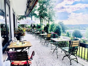An outdoor cafe with tables and chairs, a view of the countryside, and a building with large windows.