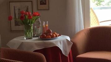 A room with a table set with fruit and a vase of red flowers. There are two framed pictures on the wall.