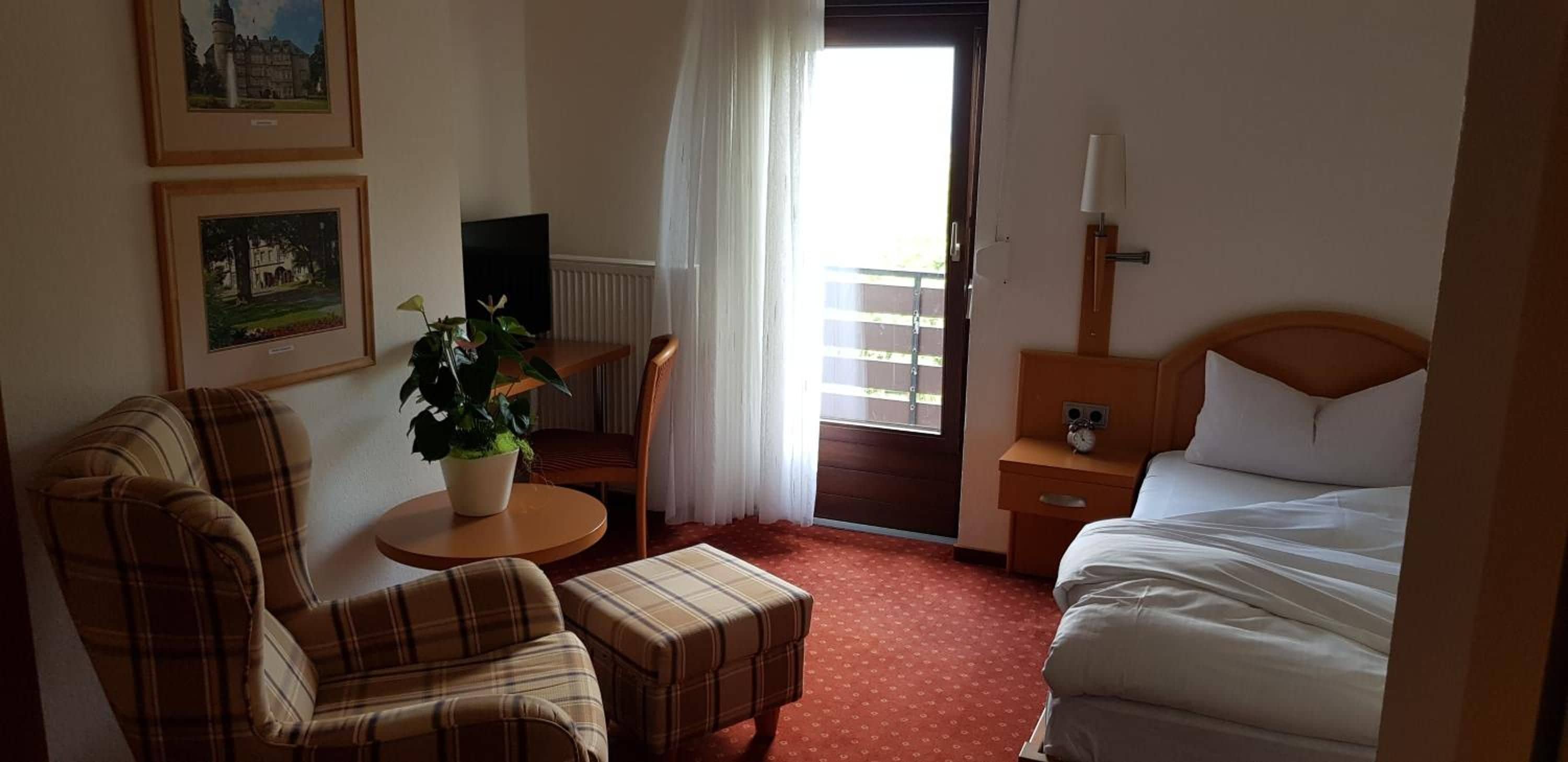 Una habitación de hotel con una cama, una silla, una mesita con una planta, una mesita de noche con una lámpara y una puerta que da a un balcón.