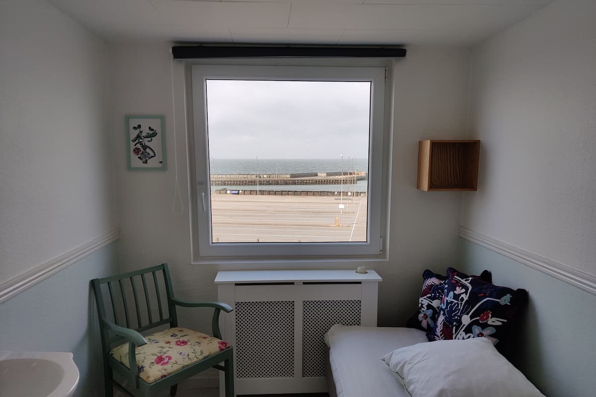 A small room with a bed, a chair, a radiator, and a window with a view of the sea and a pier.